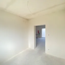 Surowe wnętrze w trakcie remontu, ściany i sufit po nałożeniu warstwy wygładzającej, widoczne kable wystające z sufitu, surowa podłoga i otwór drzwiowy prowadzący do kolejnego pomieszczenia.