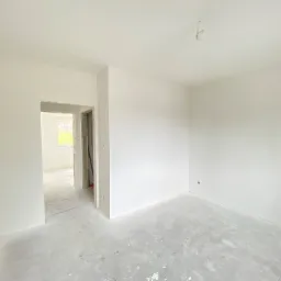 Wnętrze pokoju po wstępnym etapie prac wykończeniowych, ściany i sufit pomalowane na biało, surowa podłoga, widoczne ślady po pracach budowlanych, otwarte przejście do kolejnego pomieszczenia...
