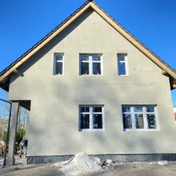 Budynek mieszkalny w trakcie ocieplania, widoczne siatka i warstwa kleju na elewacji przygotowane pod tynk zewnętrzny, z zamontowanymi oknami z folią ochronną.