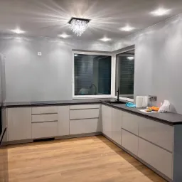 Nowoczesna kuchnia z szarymi ścianami i białymi meblami, oświetlona kryształowym żyrandolem, z widocznymi śladami prac wykończeniowych na blacie.