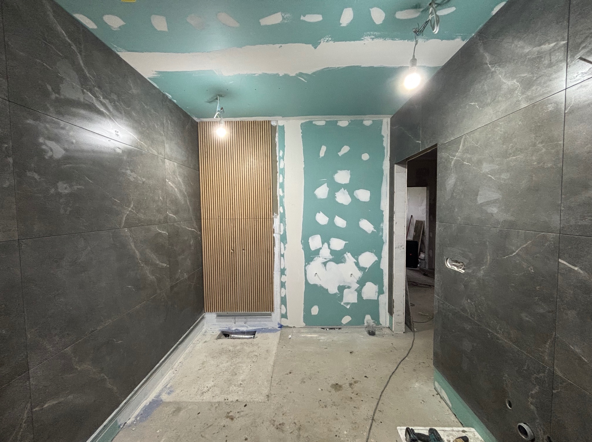 Wnętrze łazienki w trakcie remontu: ciemne płytki imitujące marmur na ścianach, pionowe drewniane lamele, surowy beton na podłodze, fragment ściany z nałożoną warstwą przygotowaną do dalszych prac...