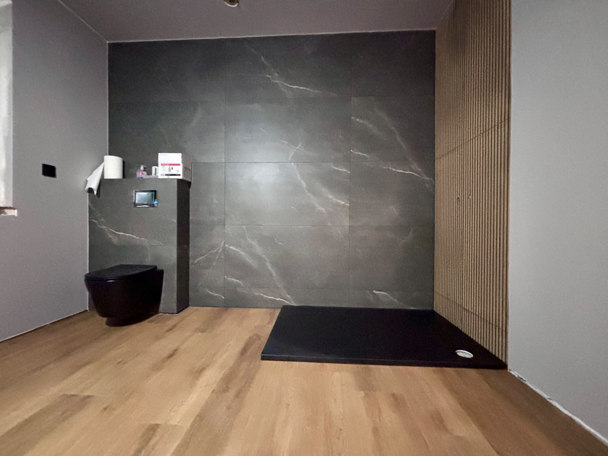 Nowoczesna łazienka z czarną miską WC podwieszaną, ciemnoszarymi płytkami imitującymi marmur, drewnianą okładziną ścienną z pionowych listew i czarnym brodzikiem bez kabiny.