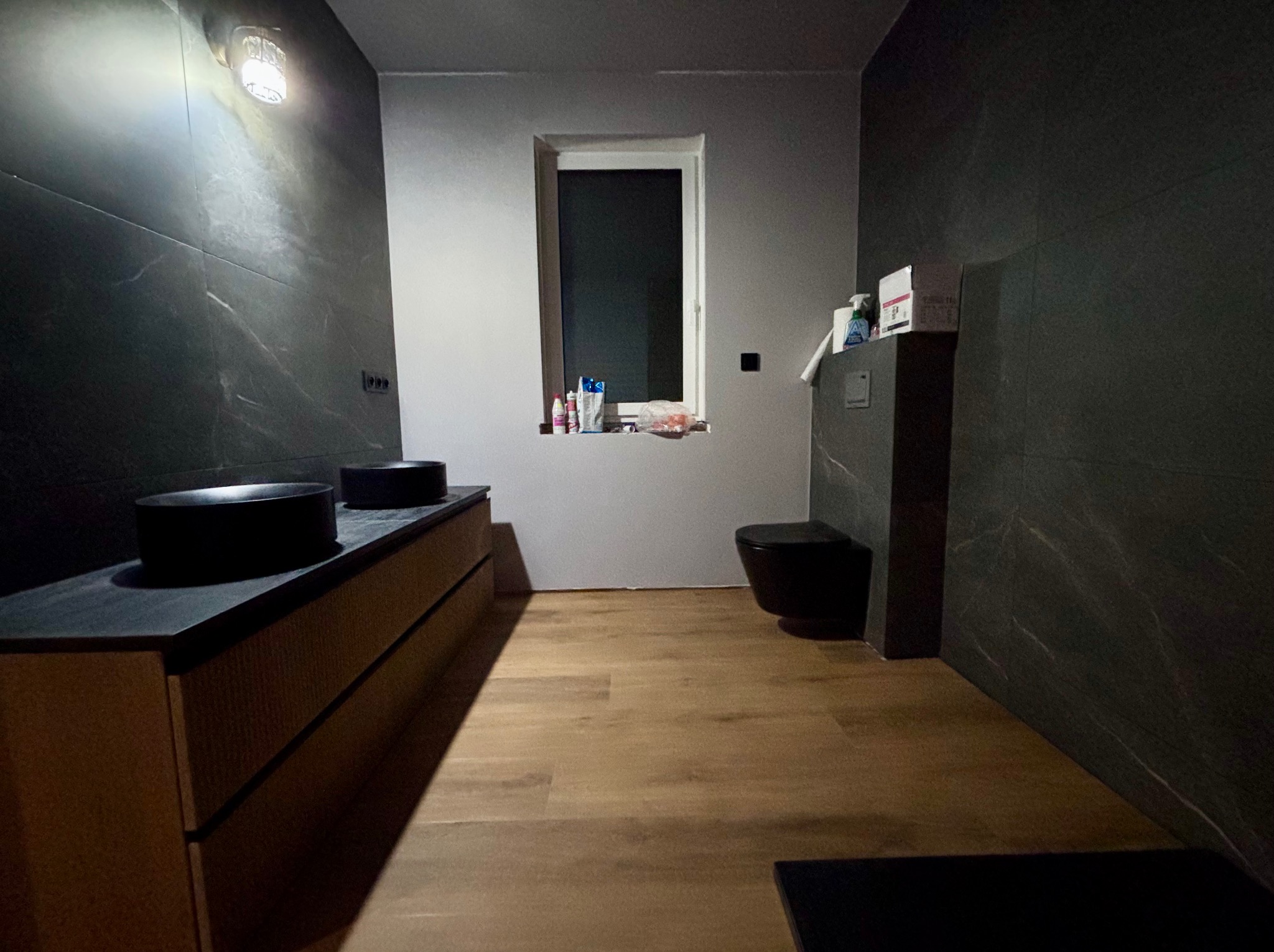 Nowoczesna łazienka z czarnymi elementami wyposażenia, drewnianą podłogą i ścianami wykończonymi płytkami imitującymi marmur. Umywalki nablatowe i wisząca toaleta.