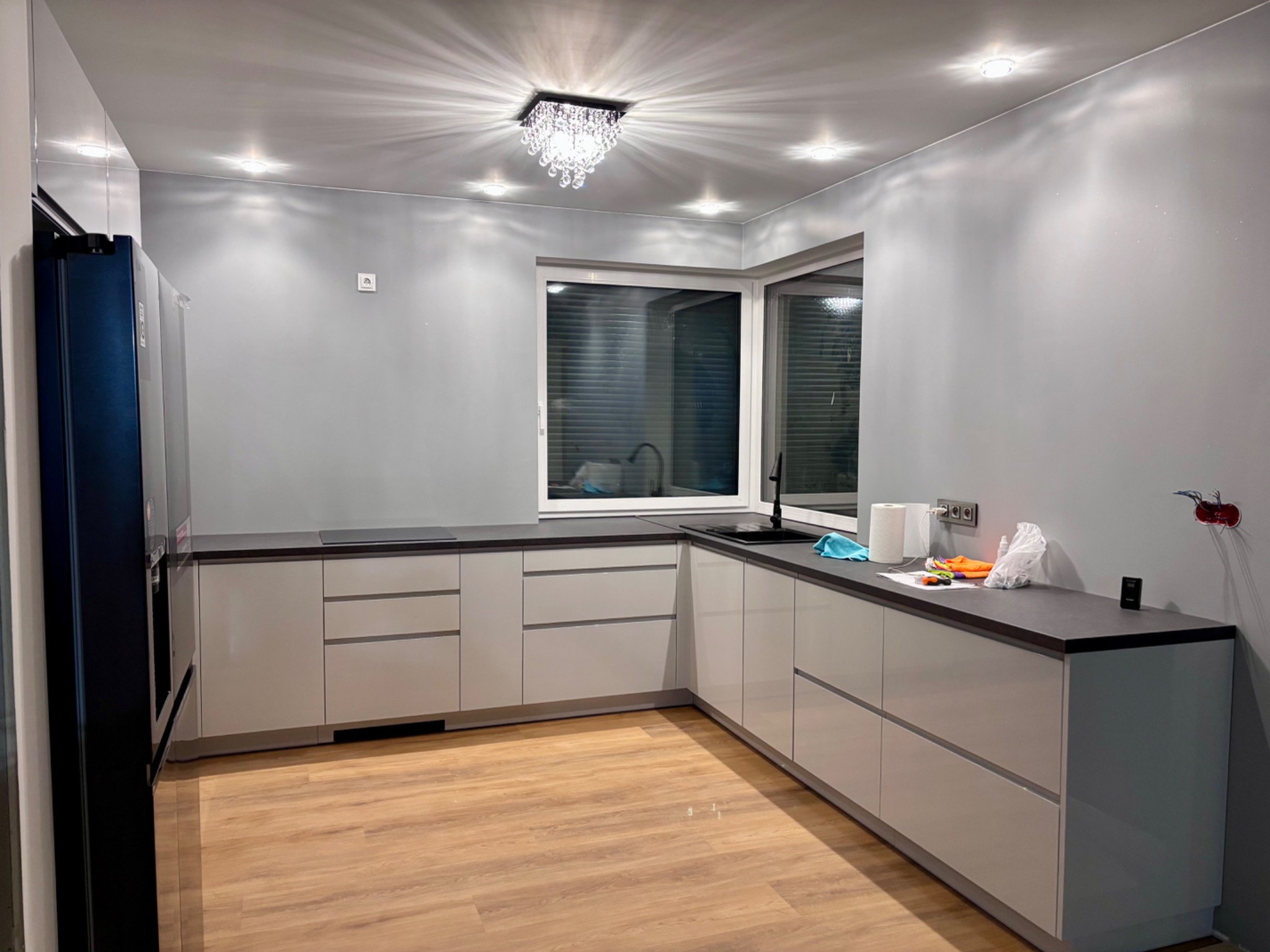 Nowoczesna kuchnia z szarymi ścianami i białymi meblami, oświetlona kryształowym żyrandolem, z widocznymi śladami prac wykończeniowych na blacie.