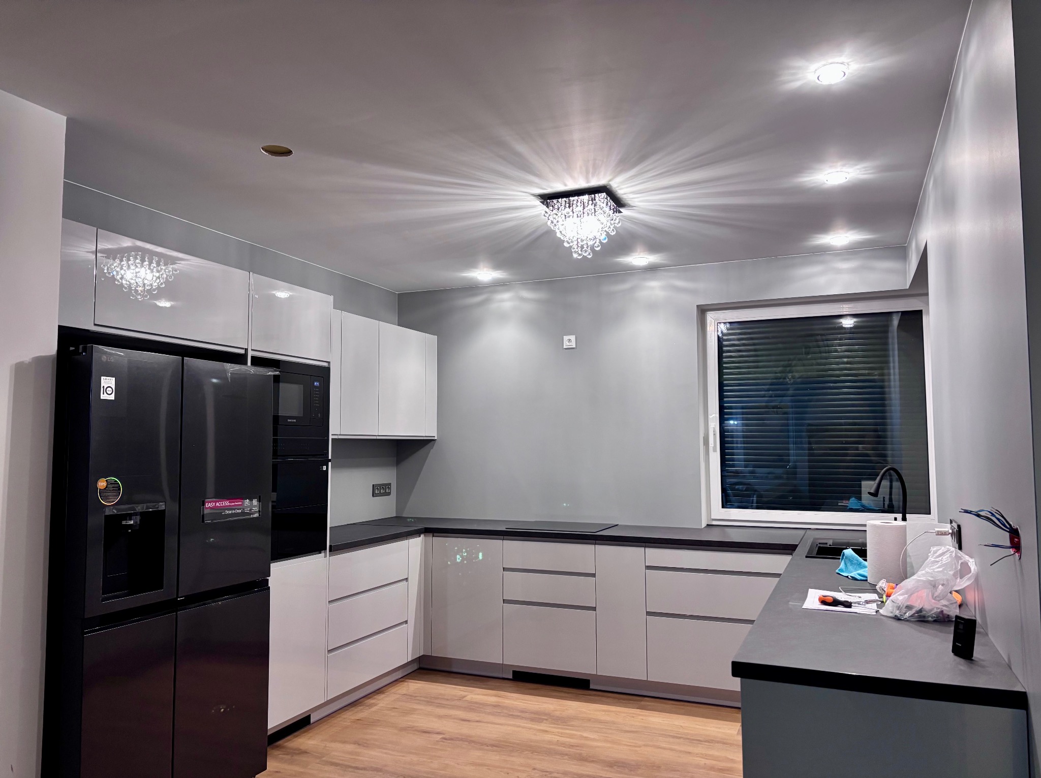 Nowoczesna kuchnia w odcieniach szarości i bieli z czarnym sprzętem AGD, widoczne niedokończone prace wykończeniowe przy zlewie.