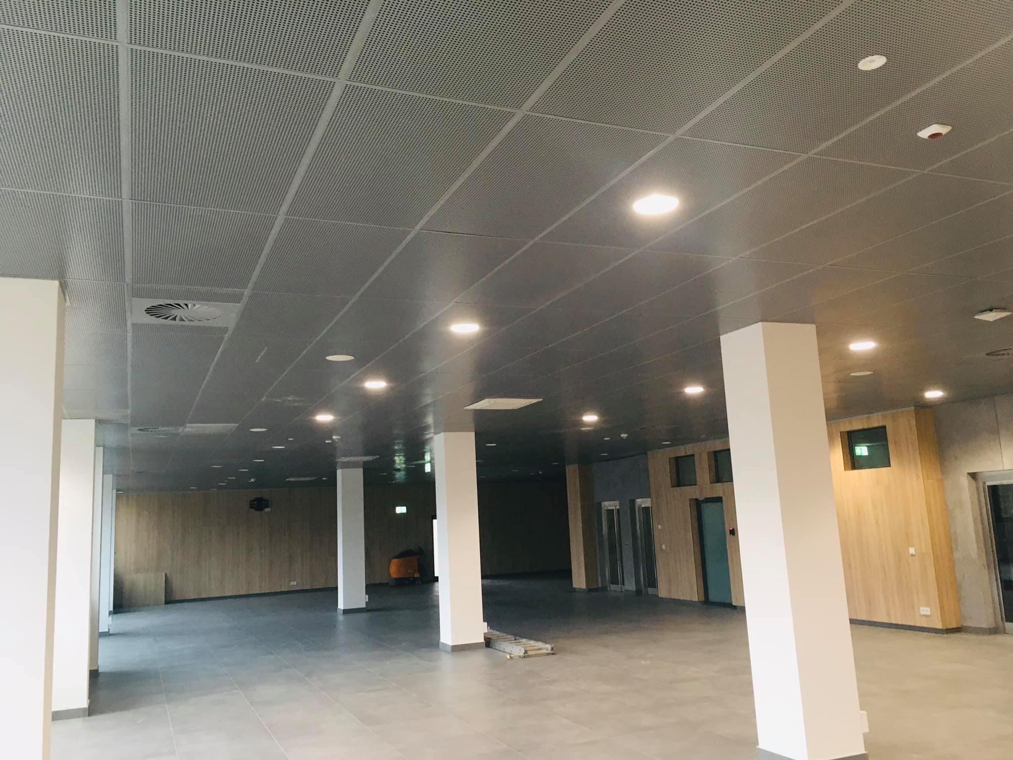 Sufit podwieszany z perforowanej płyty metalowej w dużym pomieszczeniu biurowym, widoczne białe kolumny i oświetlenie punktowe, drabina leżąca na podłodze.