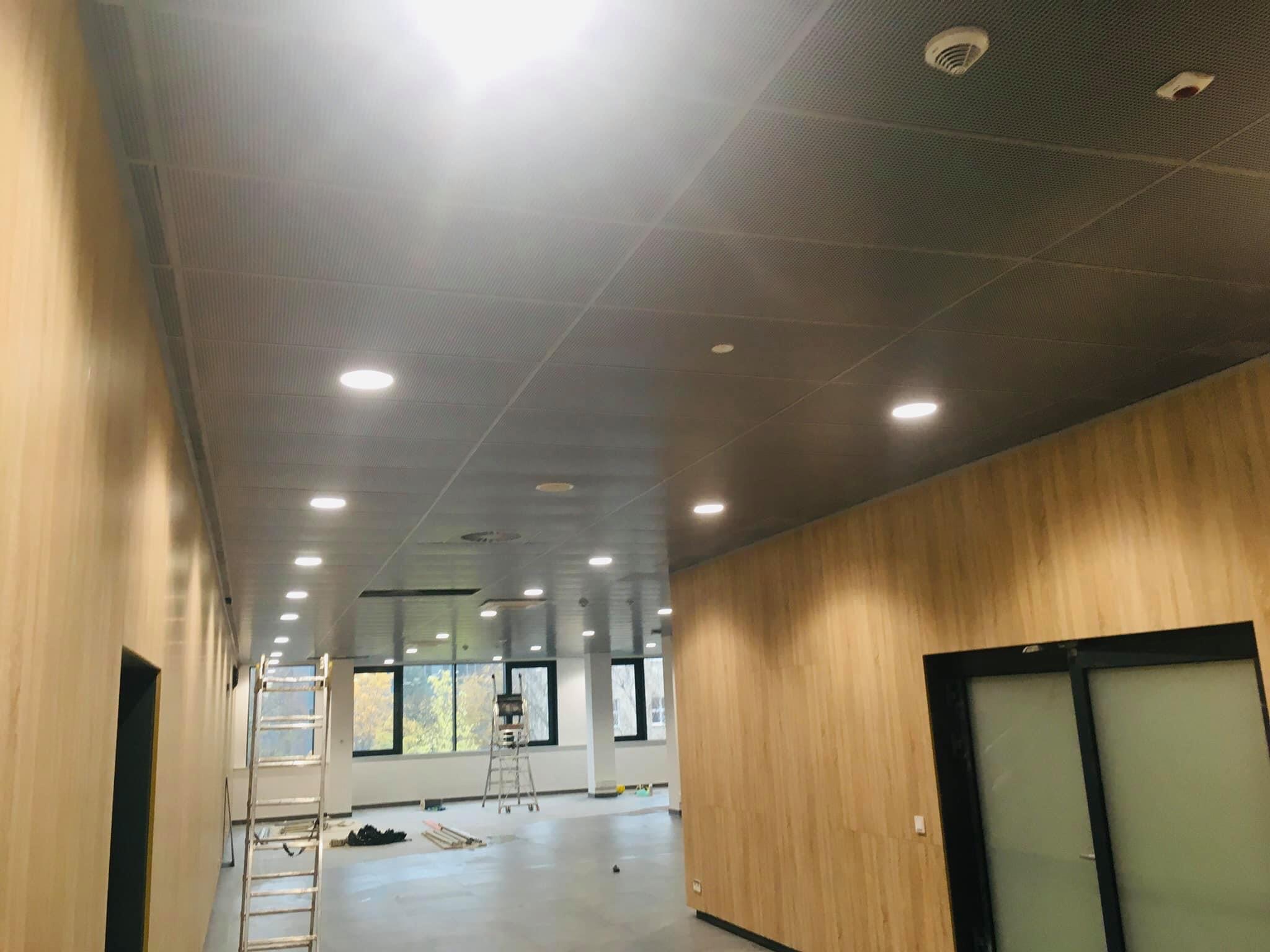 Metalowy sufit podwieszany z oświetleniem punktowym w jasnym pomieszczeniu biurowym, drabina aluminiowa ustawiona przy ścianie, widok na okno z drzewami w tle.