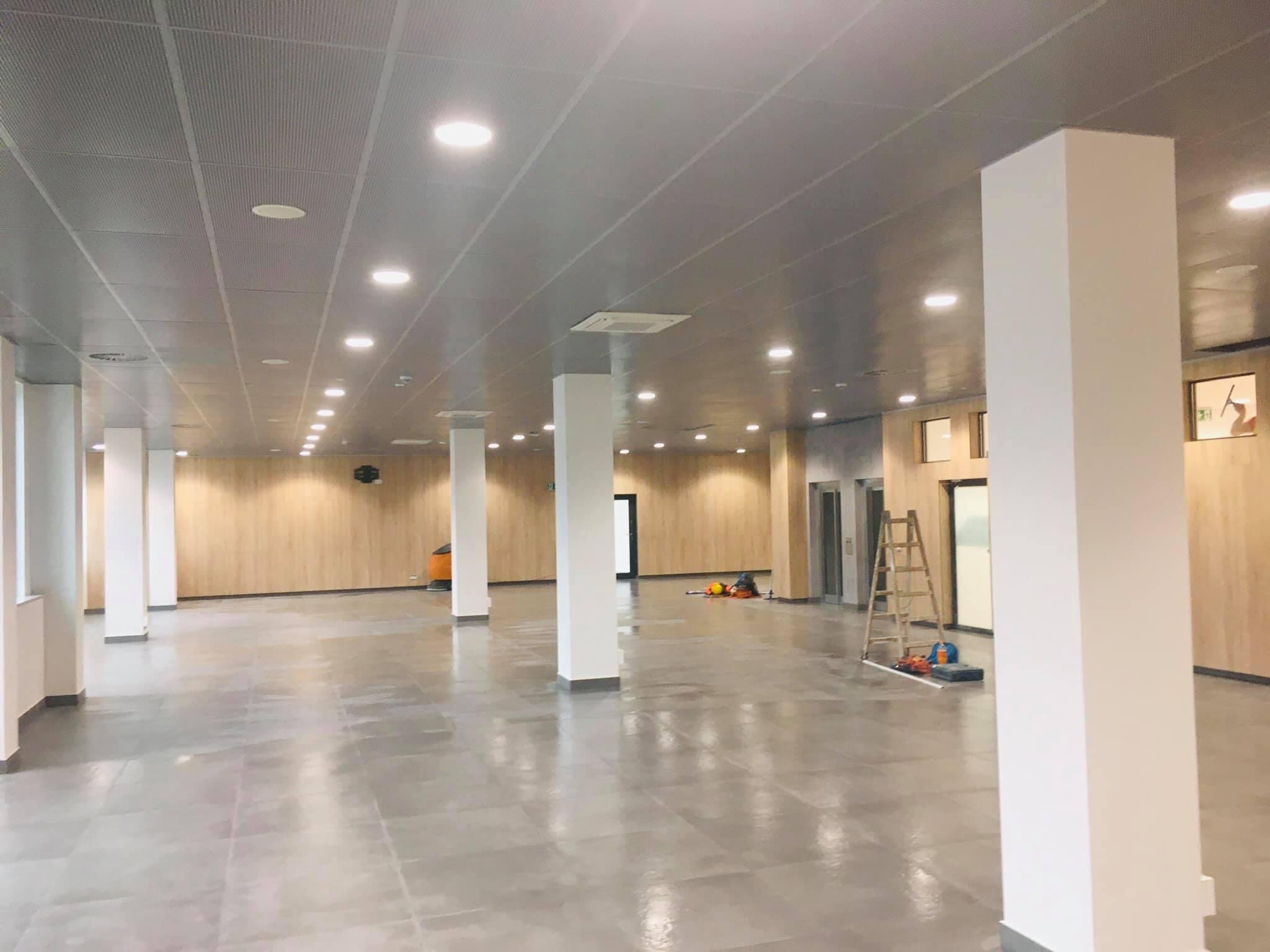 Duże, jasne pomieszczenie z metalowym, perforowanym sufitem podwieszanym, białymi kolumnami i drewnianymi panelami ściennymi w trakcie wykańczania, widoczne narzędzia i drabina.