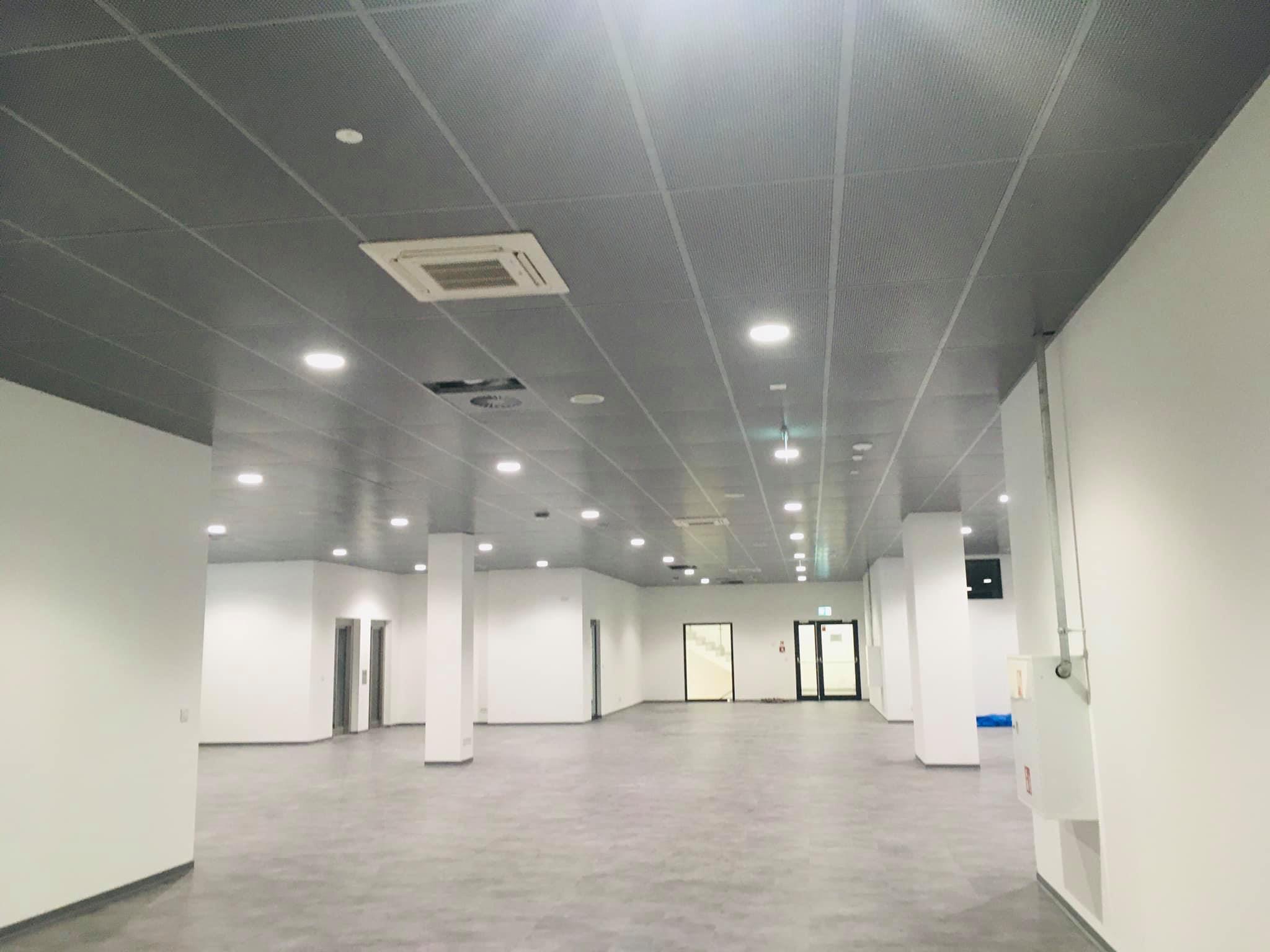 Przestronne wnętrze biurowe z szarą perforowaną konstrukcją sufitową, wbudowanymi lampami LED i centralną klimatyzacją, widoczne białe ściany i szara podłoga.