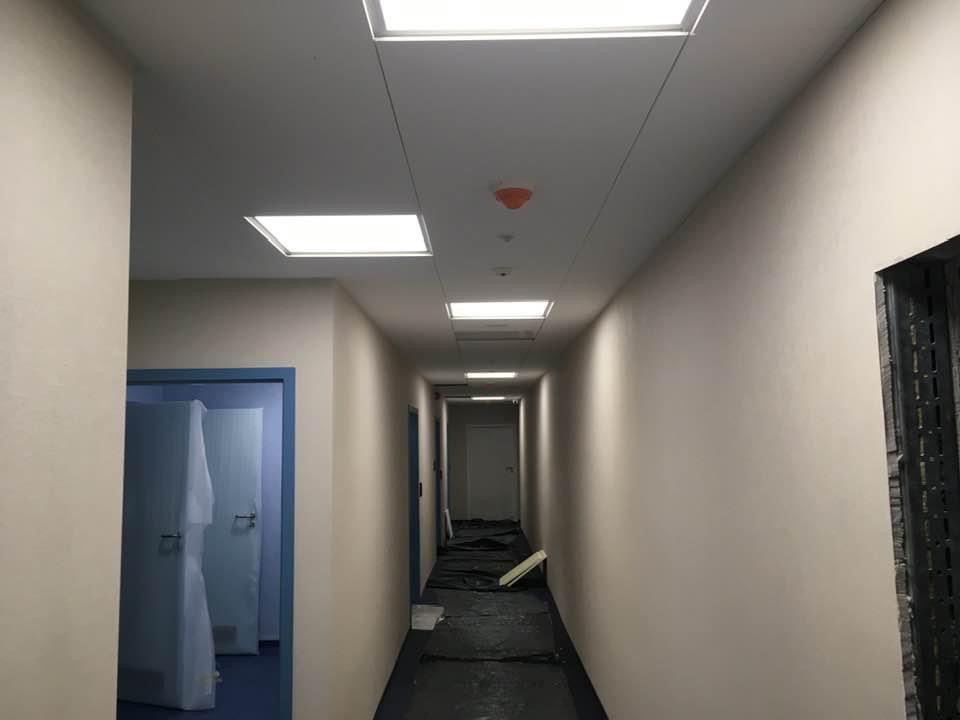 Długi korytarz z zamontowanymi panelami sufitowymi i oświetleniem LED, podłoga zabezpieczona folią malarską podczas remontu.