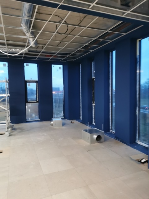 Wnętrze biurowe w trakcie montażu sufitu podwieszanego z widoczną konstrukcją metalową i instalacją wentylacyjną, ściany pomalowane na granatowo.