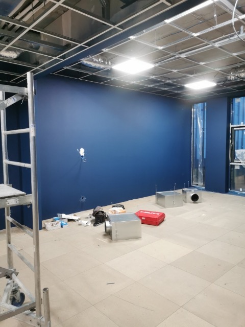 Wnętrze w trakcie remontu z pomalowaną na ciemny niebieski ścianą, widoczne metalowe elementy konstrukcji sufitu podwieszanego, rozstawione na podłodze metalowe skrzynki wentylacyjne i narzędzia...