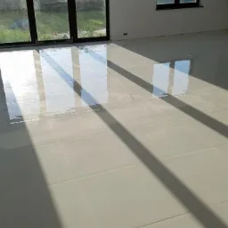 Błyszcząca, jasna podłoga z płytek w nowym domu, z wyraźnymi cieniami rzucanymi przez okno, podkreślającymi regularny wzór fug.