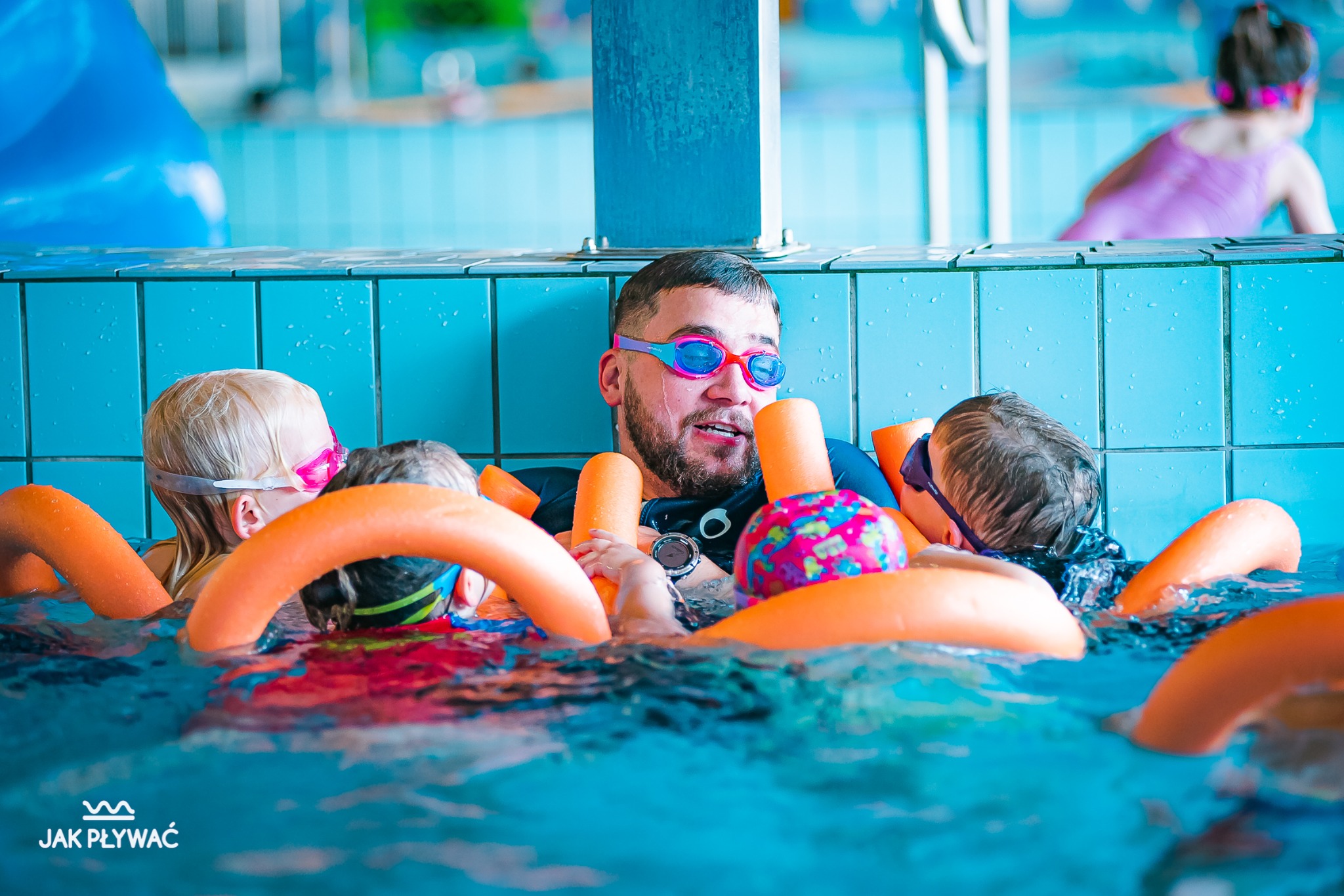 Instruktor z brodą i w okularach pływackich otoczony dziećmi z makaronami w basenie z niebieskimi kafelkami.