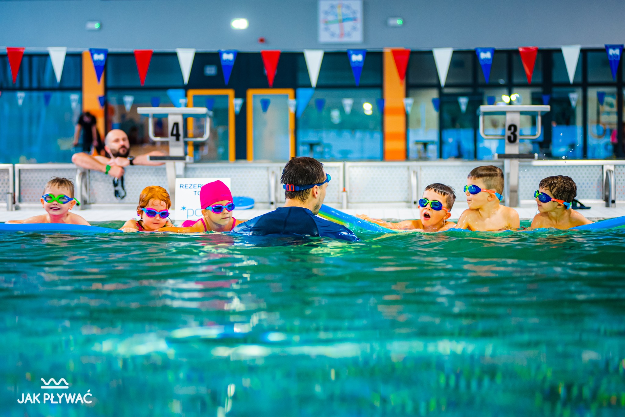 Instruktor z grupą dzieci w kolorowych goglach na basenie, podczas zajęć z pływania, w tle słupki startowe i dekoracje.