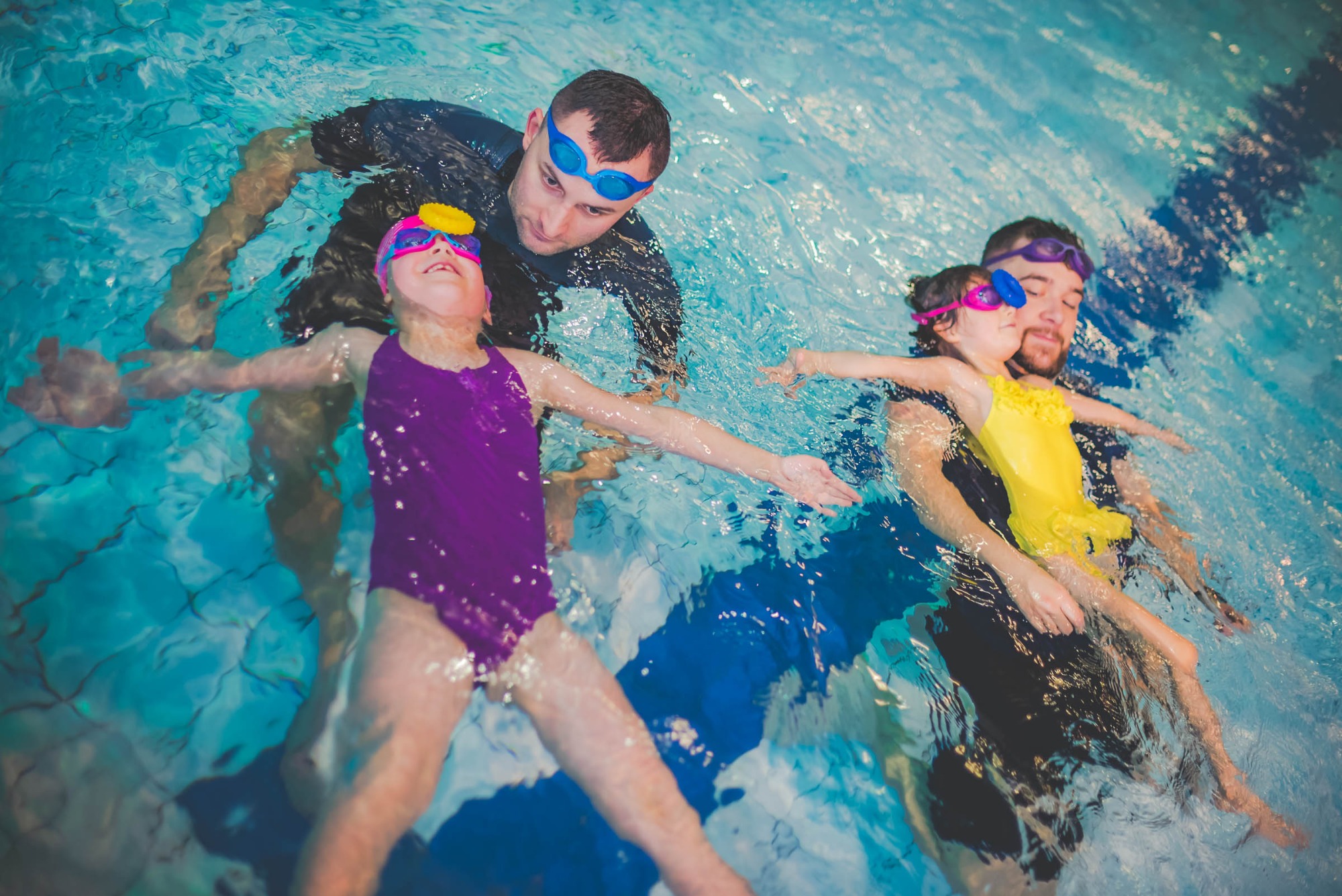 Instruktorzy z dziećmi w okularach pływackich unoszący się na wodzie w basenie. Dziewczynka w fioletowym stroju z żółtym elementem na okularach unosi się na wodzie z rozłożonymi rękami.