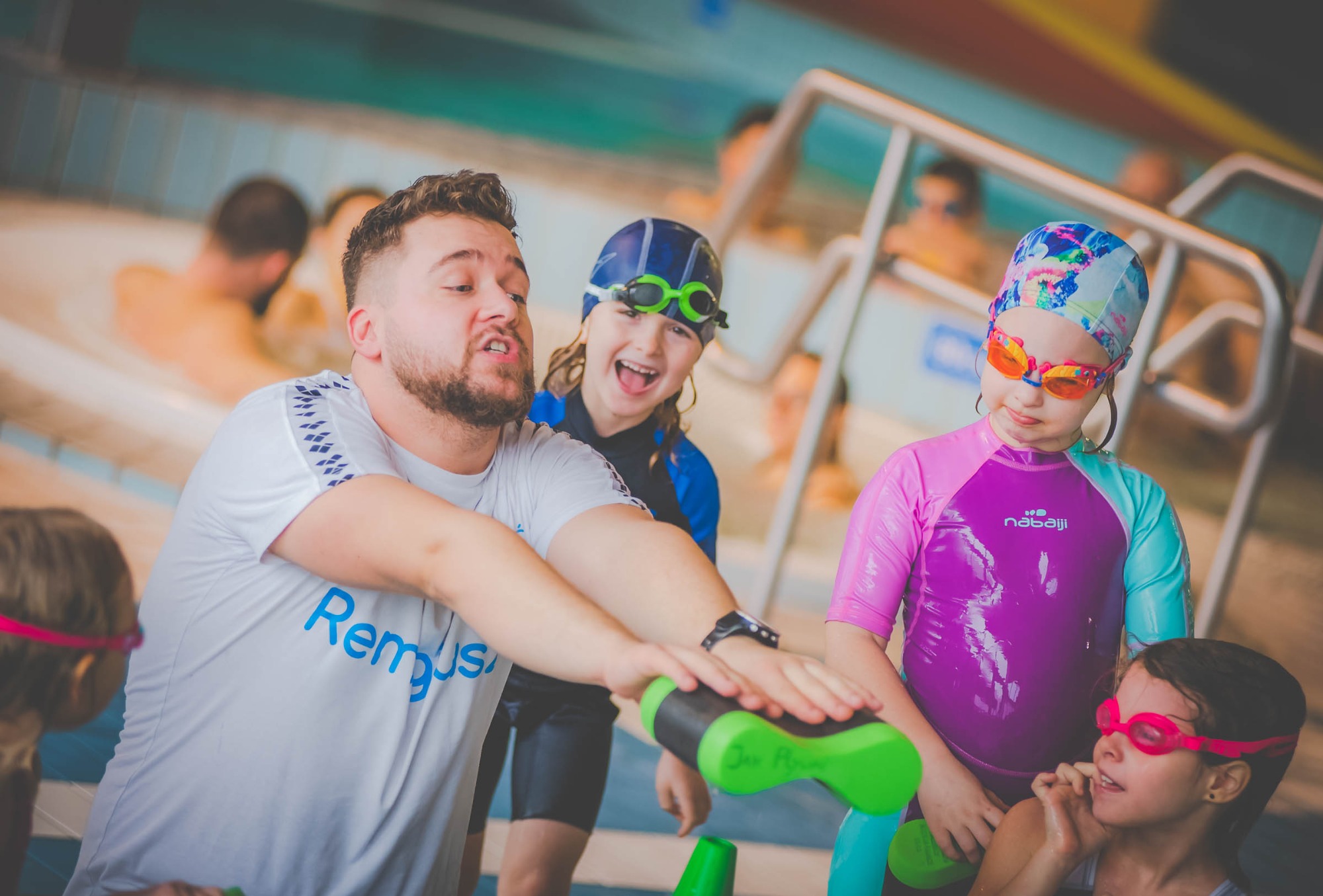 Instruktor z brodą demonstruje ćwiczenie z zielonym makaronem do pływania dzieciom w kolorowych strojach i czepkach na basenie.