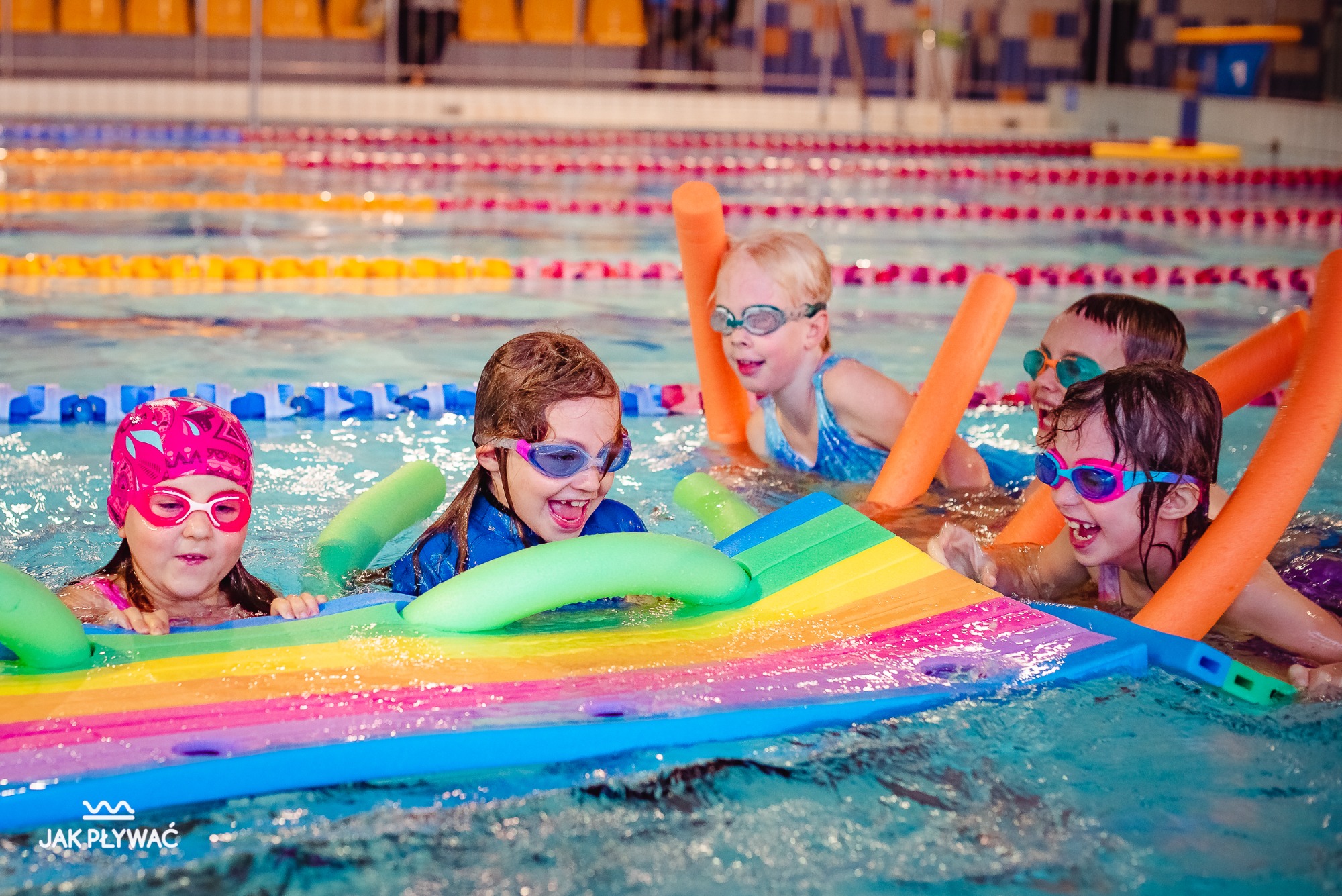 Grupa roześmianych dzieci w okularach pływackich, unoszących się na wodzie z piankowymi makaronami i kolorową deską do pływania, w tle basen z wyznaczonymi torami.
