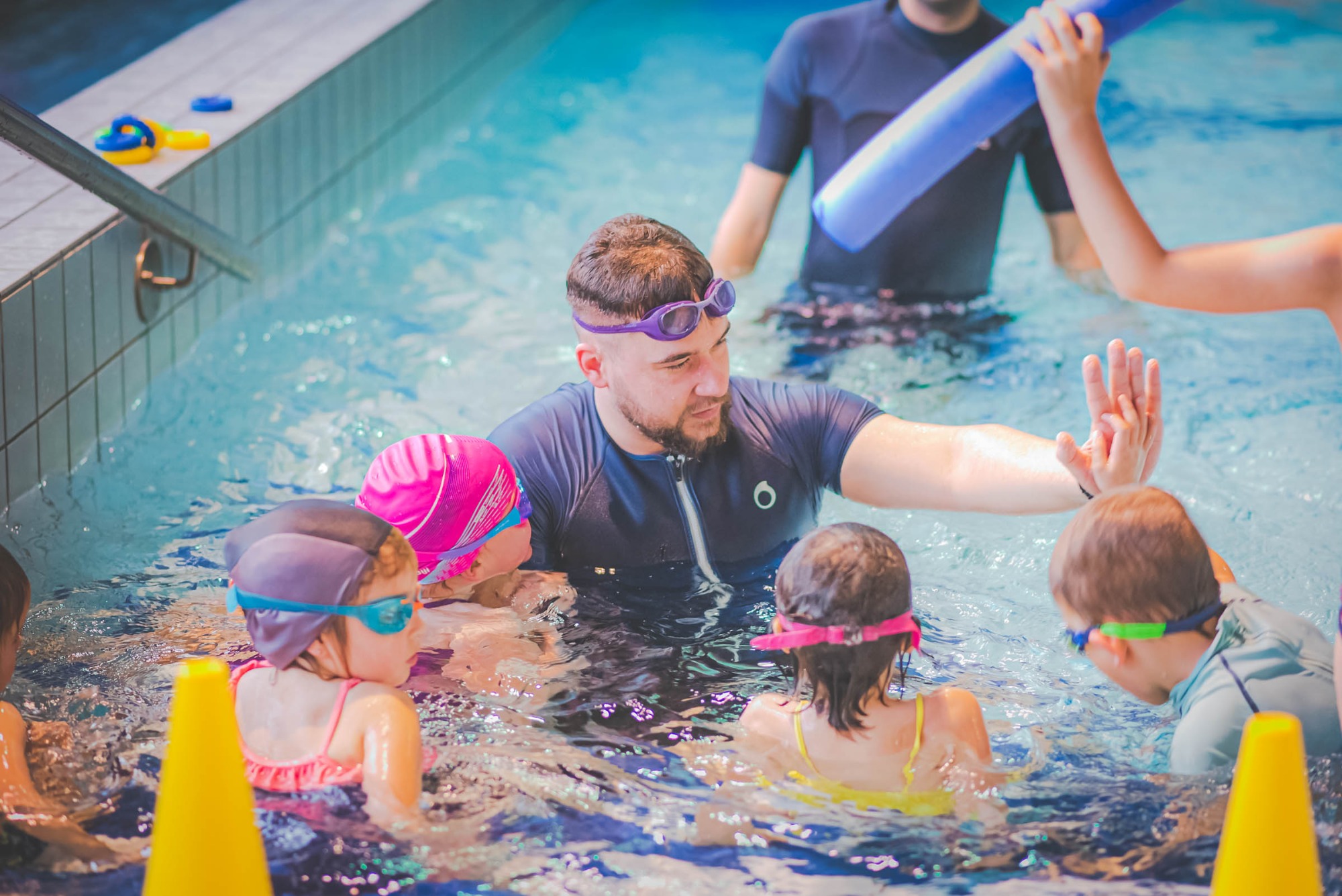 Instruktor z brodą i w fioletowych goglach prowadzi zajęcia w basenie z grupą dzieci w czepkach i goglach, używając piankowej rurki i żółtego pachołka.