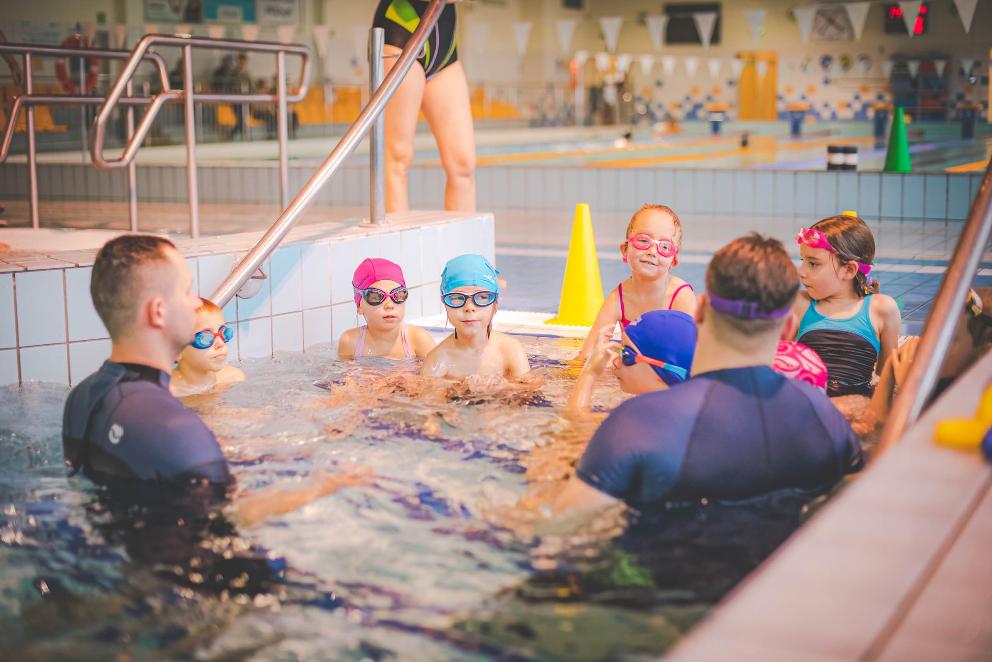 Grupa dzieci w okularach i czepkach pływackich w płytkim basenie z instruktorem, widoczne żółte pachołki na brzegu basenu.