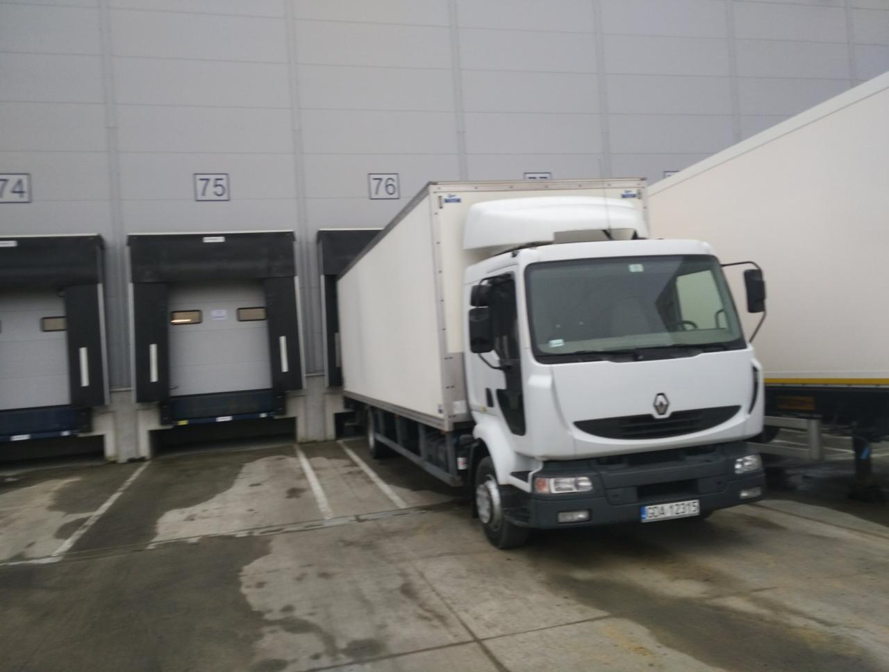 Biały samochód dostawczy Renault zaparkowany przed rampą załadunkową magazynu z numerami 74, 75 i 76.