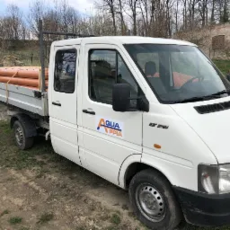 Biały samochód dostawczy z pomarańczowymi rurami na pace, zaparkowany na trawiastym terenie, widoczne logo firmy Aqua PPIA na drzwiach.