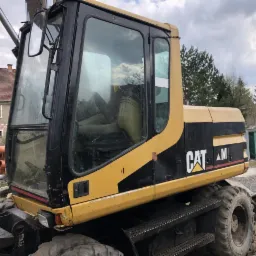 Żółto-czarna koparka kołowa marki CAT na placu budowy, widoczne ceramiczne rury w tle, pochmurne niebo.