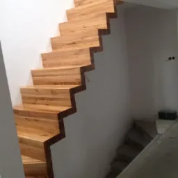 Drewniane schody bez poręczy, o jasnym odcieniu drewna, zamontowane w surowym wnętrzu z widocznymi betonowymi schodami w tle.