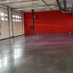 Wnętrze hali garażowej z betonową posadzką, czerwonymi ścianami i białymi bramami segmentowymi z oknami, widoczne systemy wentylacyjne pod sufitem.