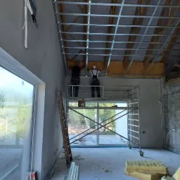 Prace wykończeniowe poddasza: dwaj mężczyźni na rusztowaniu montują izolację z wełny mineralnej między metalowymi profilami sufitowymi, widoczne elementy drewnianej konstrukcji dachu oraz fragment...