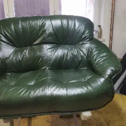 Odnowiona dwuosobowa sofa w ciemnozielonej skórze, widoczna na tle okna i materiałów tapicerskich, z widocznymi śladami pracy w warsztacie.