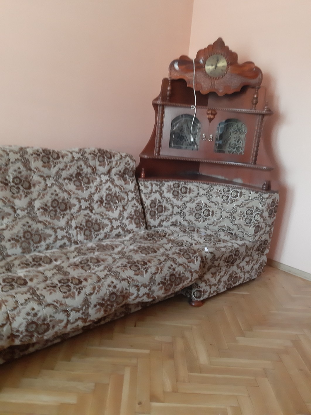 Rozłożona sofa narożna z wzorem kwiatowym w odcieniach brązu i beżu, stojąca obok drewnianej szafki narożnej z zegarem, na tle bladoróżowej ściany i parkietu.