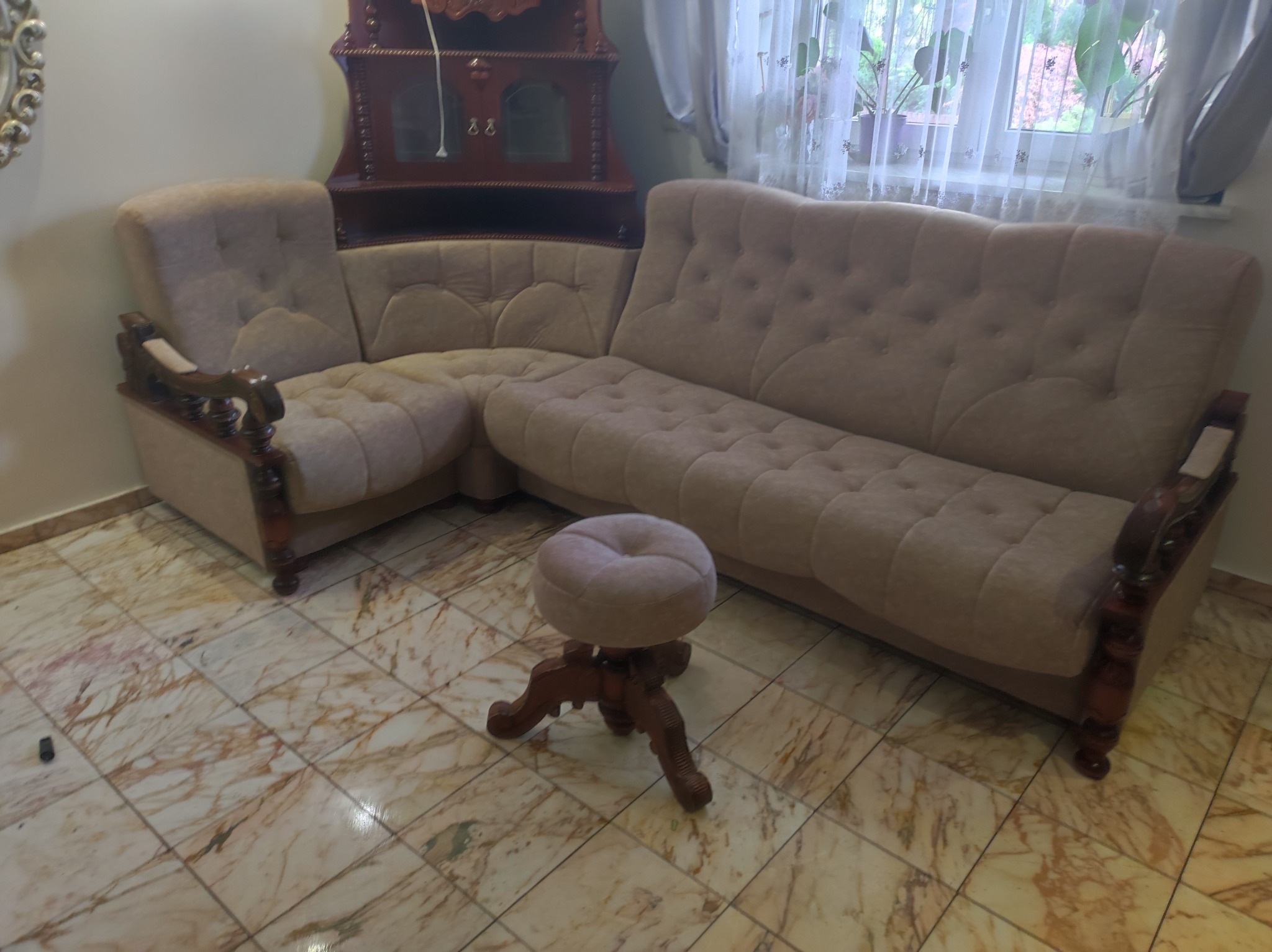 Beżowy narożnik z drewnianymi zdobieniami i pufa na trójnożnej podstawie w pokoju z marmurową podłogą i drewnianym kredensem w tle.