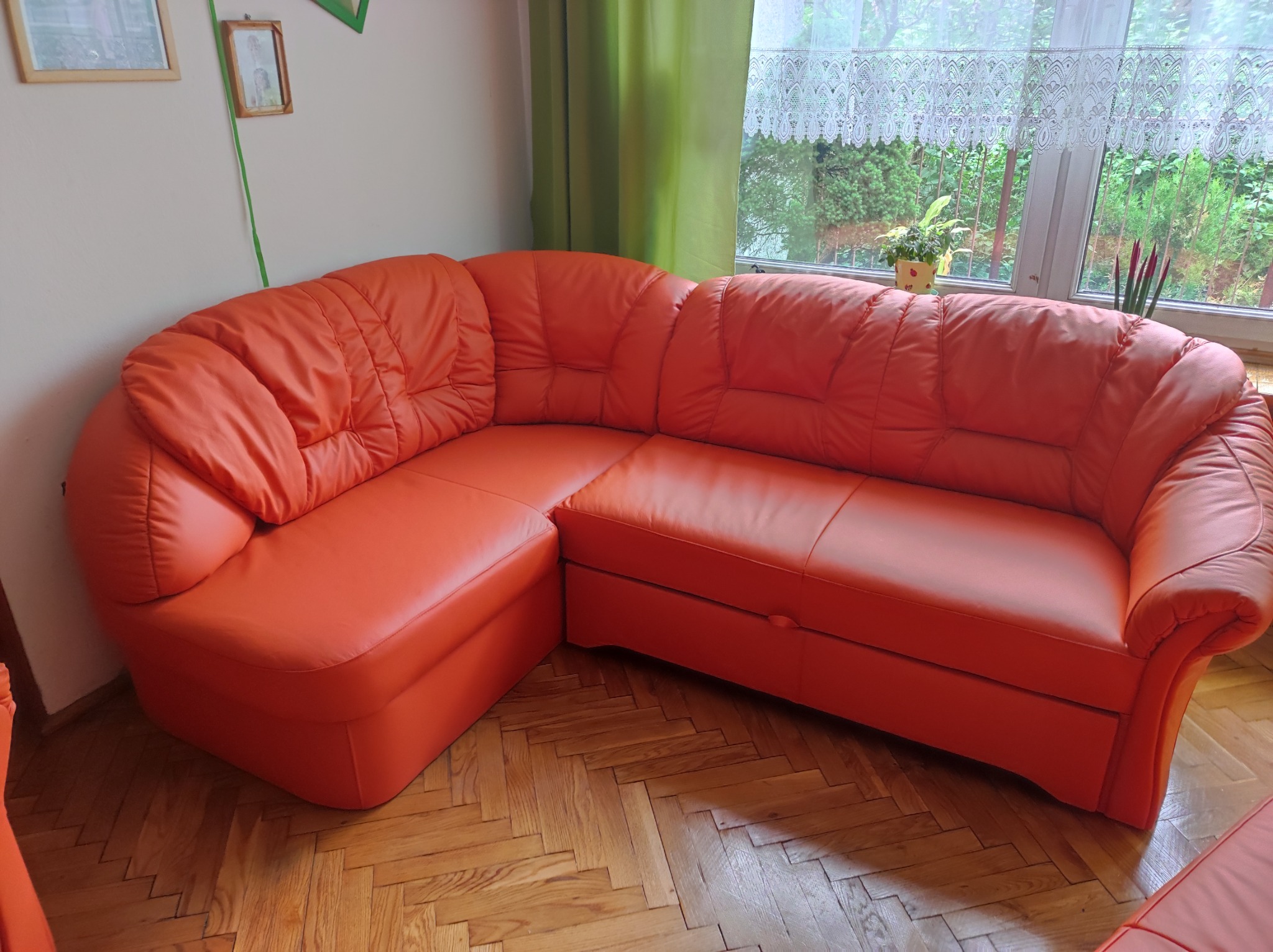 Pomarańczowy, skórzany narożnik z pikowanymi poduchami stoi na parkiecie w jodełkę, widok z boku, w tle okno z firanką i zielenią za oknem.