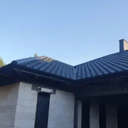 Niski kąt widzenia na dom w trakcie budowy z nowym, ciemnoszarym dachem z blachodachówki, widoczny komin i fragment ściany z białych bloczków oraz częściowo zamontowane rolety.