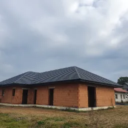 Nowo wybudowany dom z czerwonej cegły z grafitowym dachem z blachodach&oacute;wki, dekarz na rusztowaniu montuje obr&oacute;bki blacharskie, pochmurne niebo w tle.