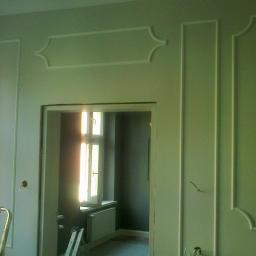 Andrzej Ciesiński - Ściana w kolorze pistacjowym z dekoracyjnymi listwami sztukateryjnymi, widok na sąsiednie pomieszczenie przez otwór drzwiowy, drabina aluminiowa oparta o ścianę.