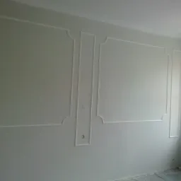 Ściana w pokoju z dekoracyjnymi listwami ściennymi w kolorze białym, widoczne przygotowania do malowania, zabezpieczona folia na podłodze i butelki na parapecie.