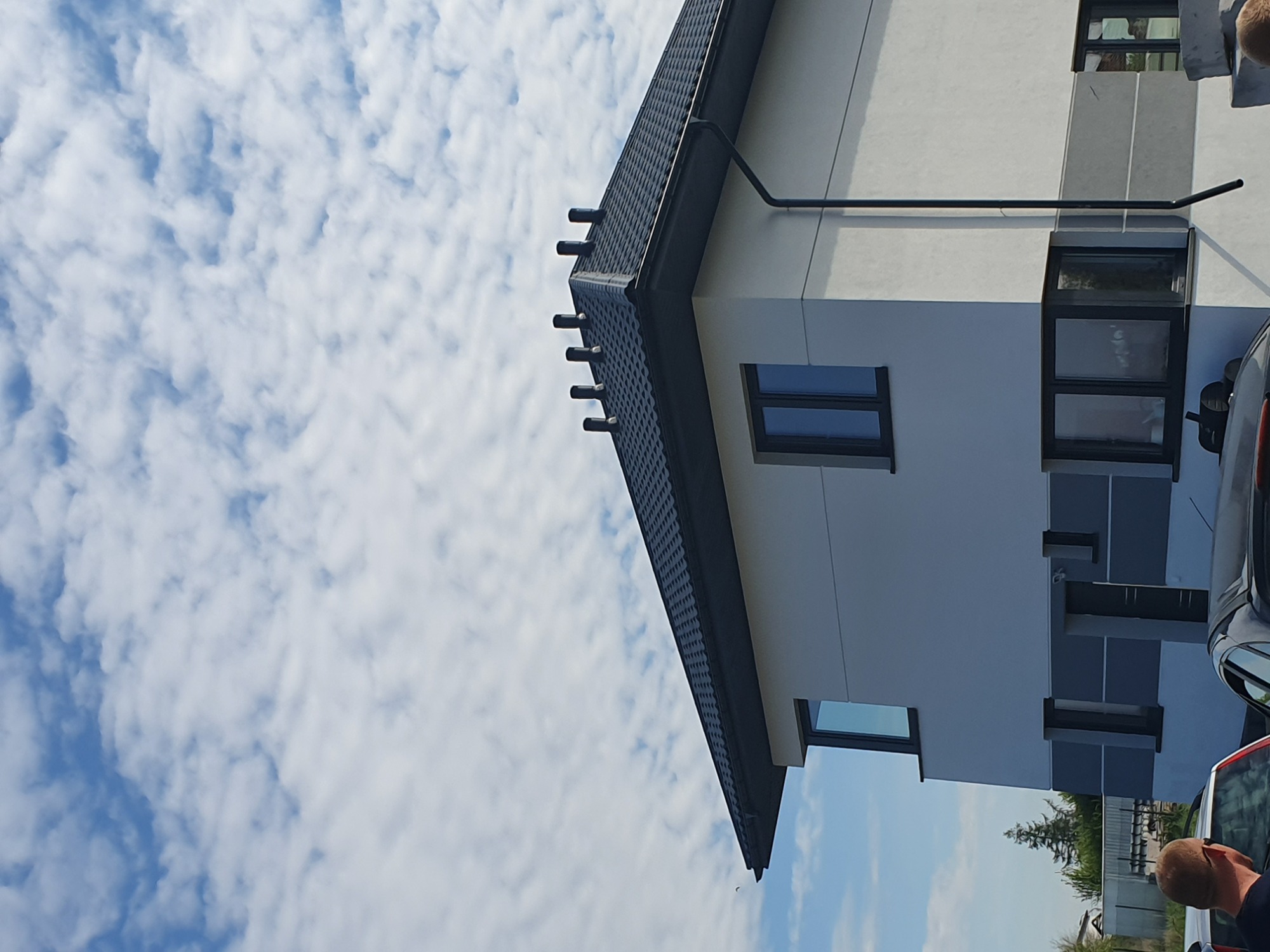 Fragment nowoczesnego domu z ciemnoszarą, metalową dachówką na tle błękitnego nieba z obłokami.