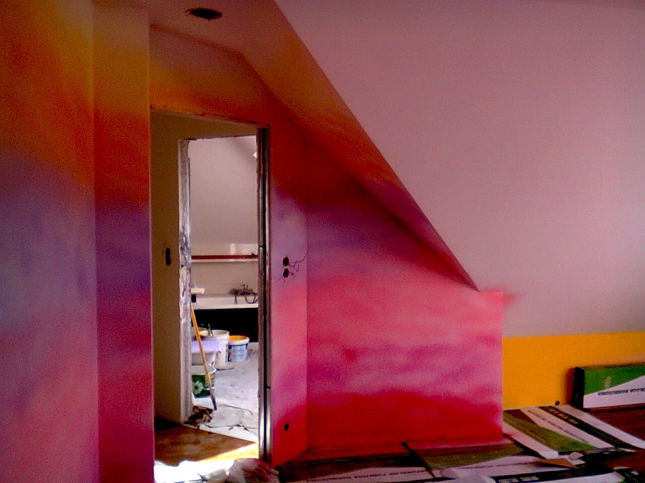 Ściana poddasza w trakcie malowania na różowo, widoczne przejścia kolorów i niedomalowane fragmenty, w tle otwarte drzwi do łazienki z wiadrami i narzędziami