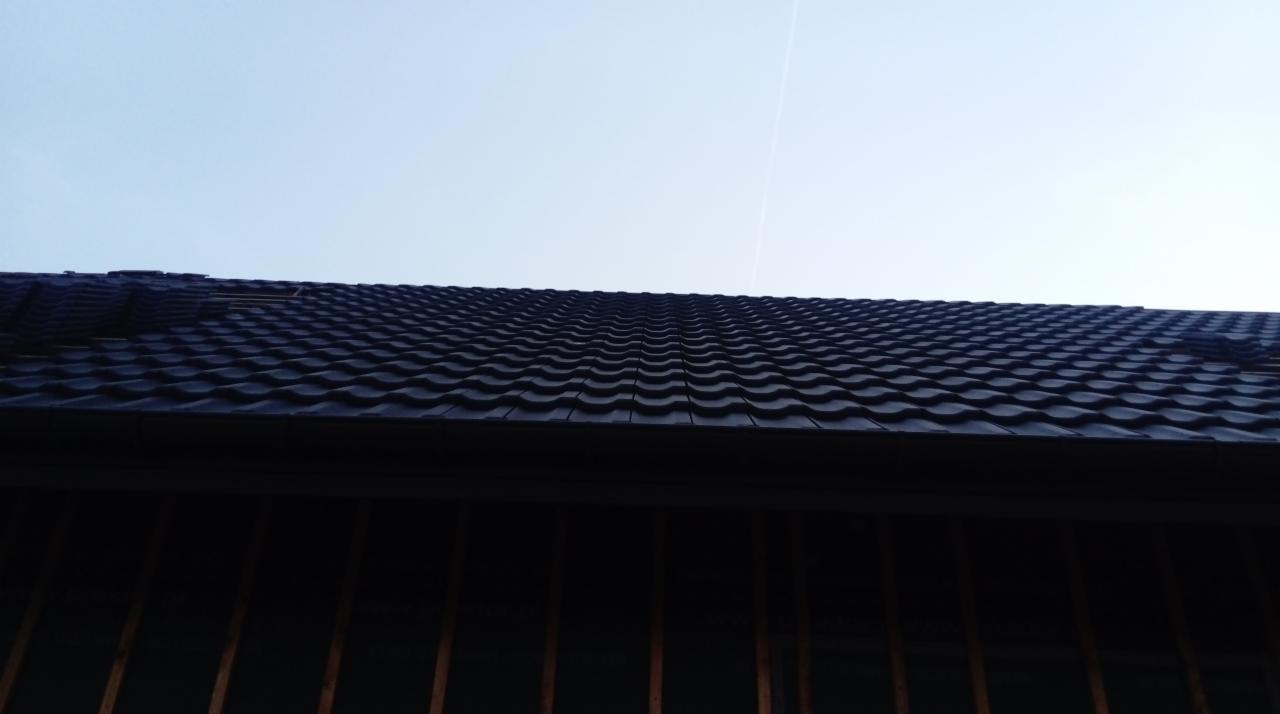 Ciemna, metalowa dachówka modułowa na nowym dachu, widok z dołu na tle jasnego nieba.