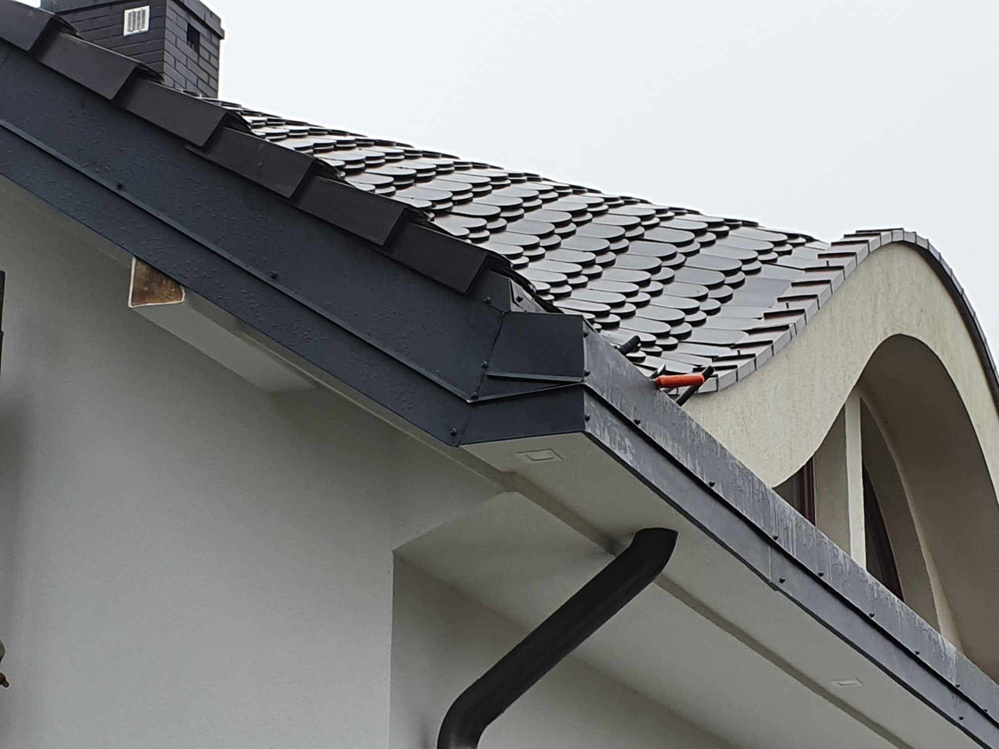 Fragment dachu z czarnej dachówki karpiówki, widoczny komin z ciemnej cegły, orynnowanie i fragment elewacji budynku z oknem w kształcie łuku.