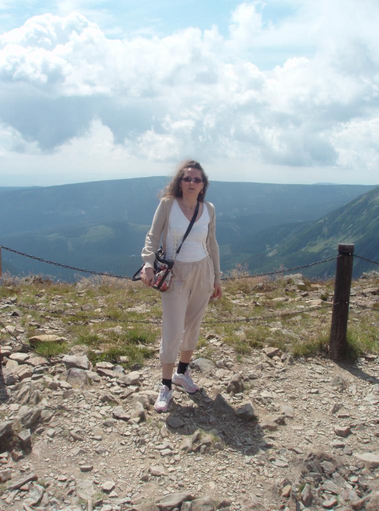 Kobieta w okularach przeciwsłonecznych, ubrana w jasne spodnie i bluzkę, stoi na kamienistym zboczu góry z widokiem na odległe szczyty i chmurne niebo.