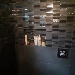 Ciemna łazienka z mozaiką na ścianie, p&oacute;łka z kosmetykami i czarny panel sterowania prysznicem.