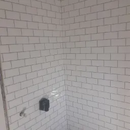 Ściana łazienki wyłożona białymi, prostokątnymi płytkami typu metro z szarą fugą, widoczne przyłącza wodne i zakryty zawór, narożnik pomieszczenia