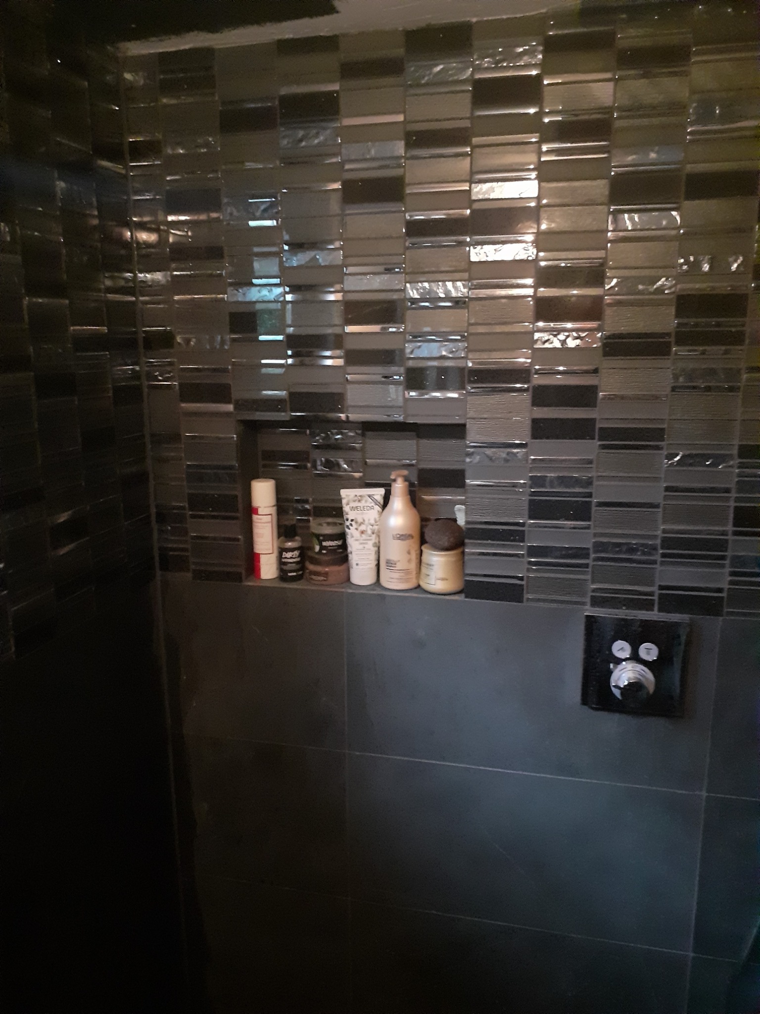 Ciemna łazienka z mozaiką na ścianie, półka z kosmetykami i czarny panel sterowania prysznicem.