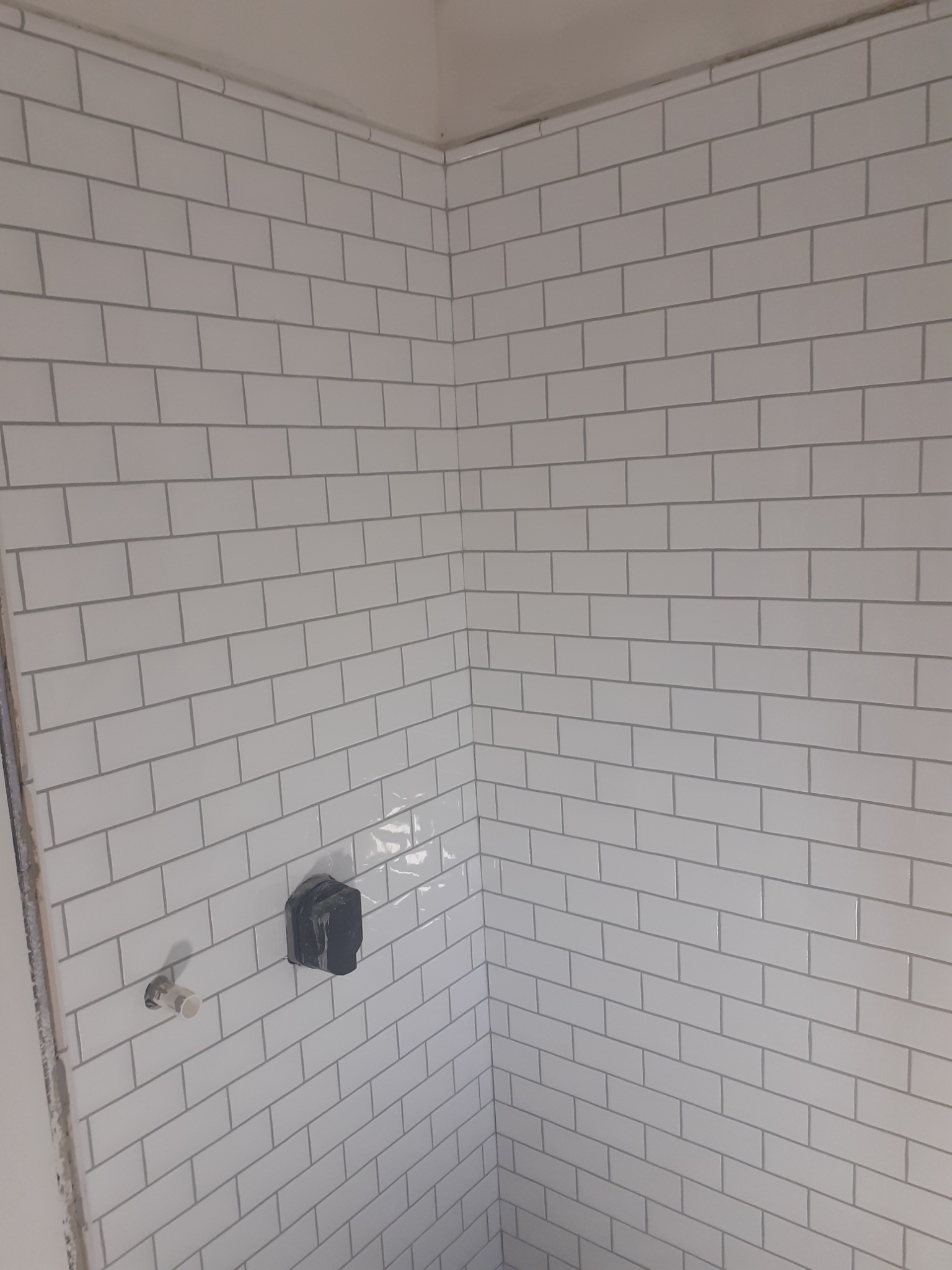 Ściana łazienki wyłożona białymi, prostokątnymi płytkami typu metro z szarą fugą, widoczne przyłącza wodne i zakryty zawór, narożnik pomieszczenia