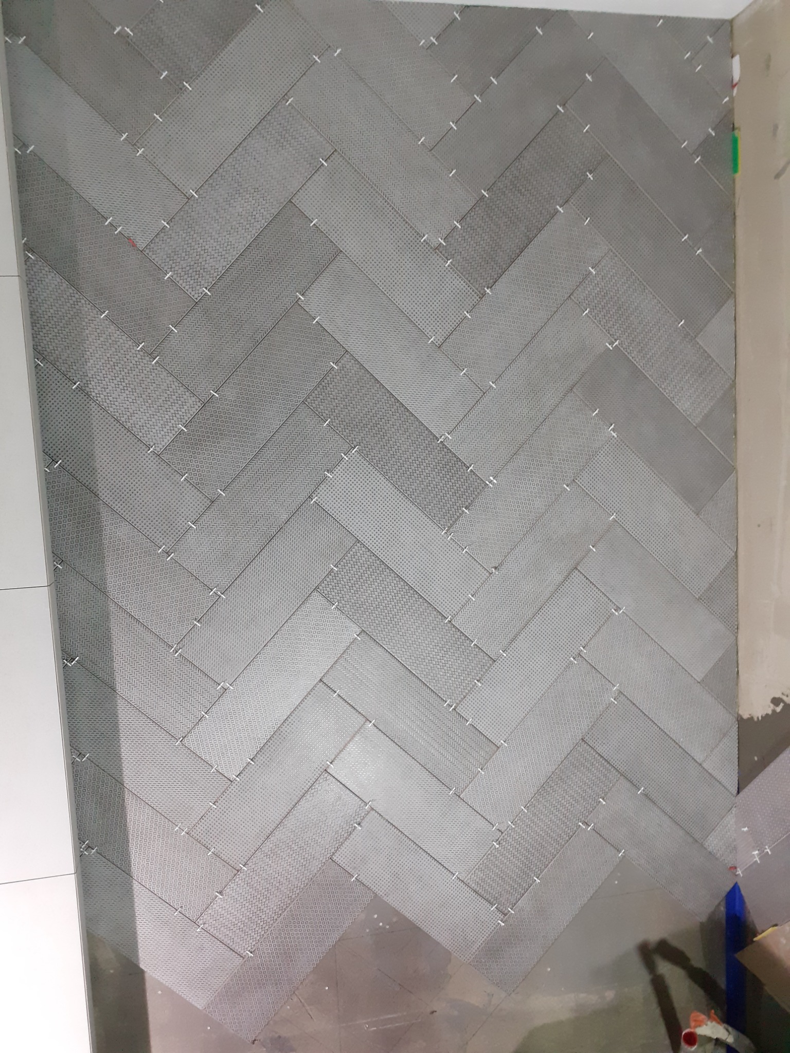 Ułożona szara mozaika w jodełkę z białymi krzyżykami dystansowymi, widoczne niedoskonałości podłoża i fragment ściany w trakcie prac.