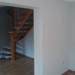 Świeżo pomalowane wnętrze z drewnianymi schodami zabezpieczonymi folią malarską, widoczna podłoga z paneli w kolorze drewna i białe ściany z widocznymi gniazdkami elektrycznymi.