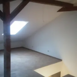 Poddasze w trakcie adaptacji z widoczną drewnianą konstrukcją nośną, oknem dachowym i otworem w podłodze, częściowo wykończone ściany, na podłodze widoczne narzędzia i materiały budowlane.